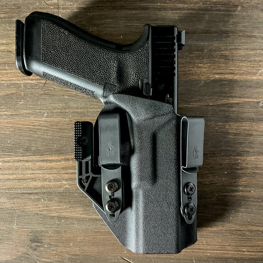 A black polymer IWB (Inside the Waistband) holster holding a handgun, photographed from a top-down perspective on a dark wood surface.